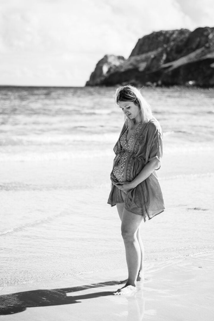 Photo en noir et blanc d'une femme enceinte sur une plage qui touche son ventre rond